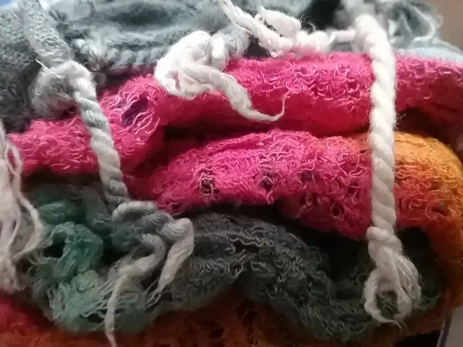 A pile of soft woollens in pink, rust and green with loose fringe threads across the top